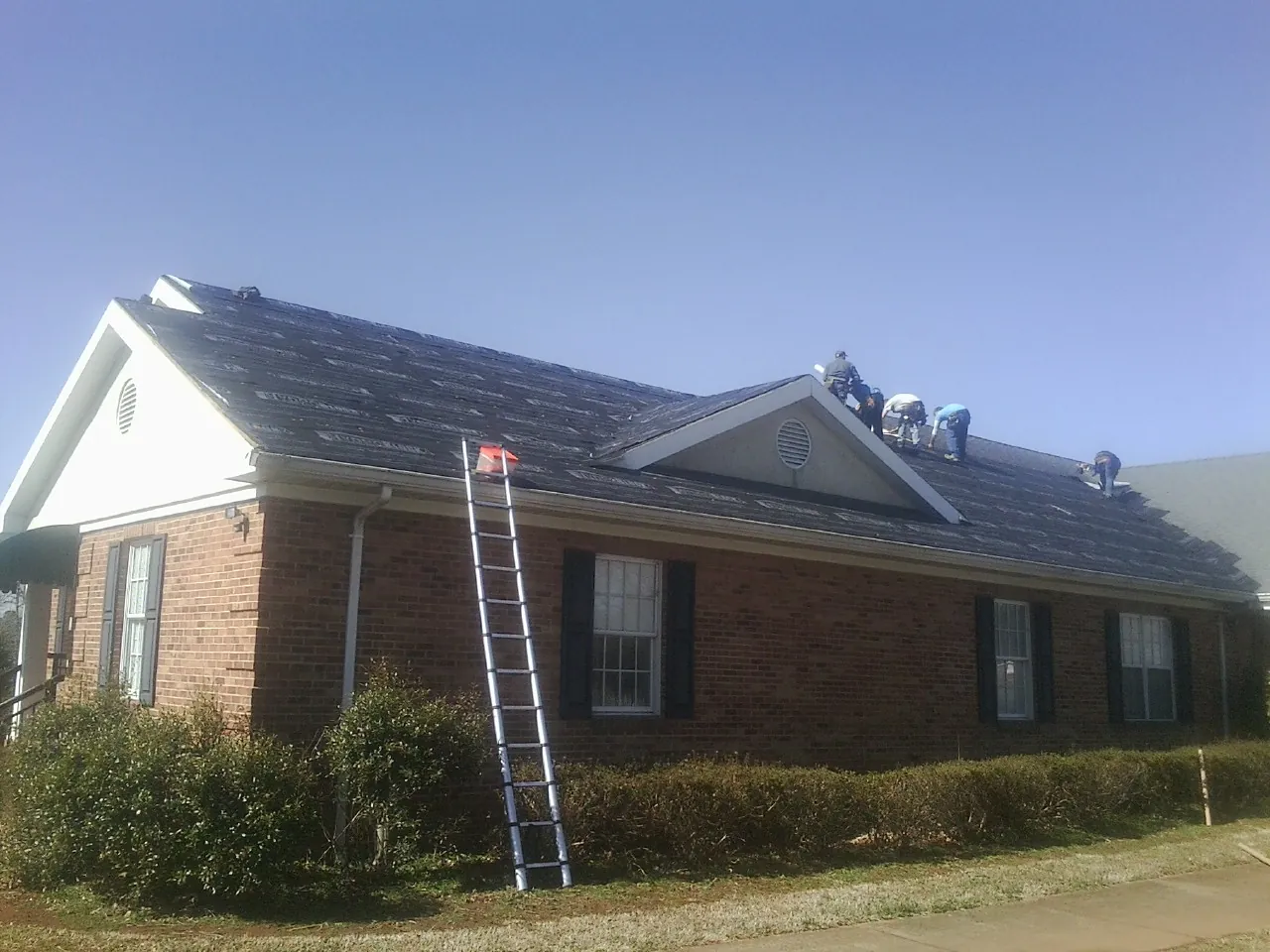 Crew tearing shingles off a brick ranch roof with a ladder set — Emergency Roof Repair work by our crew in Hamlet