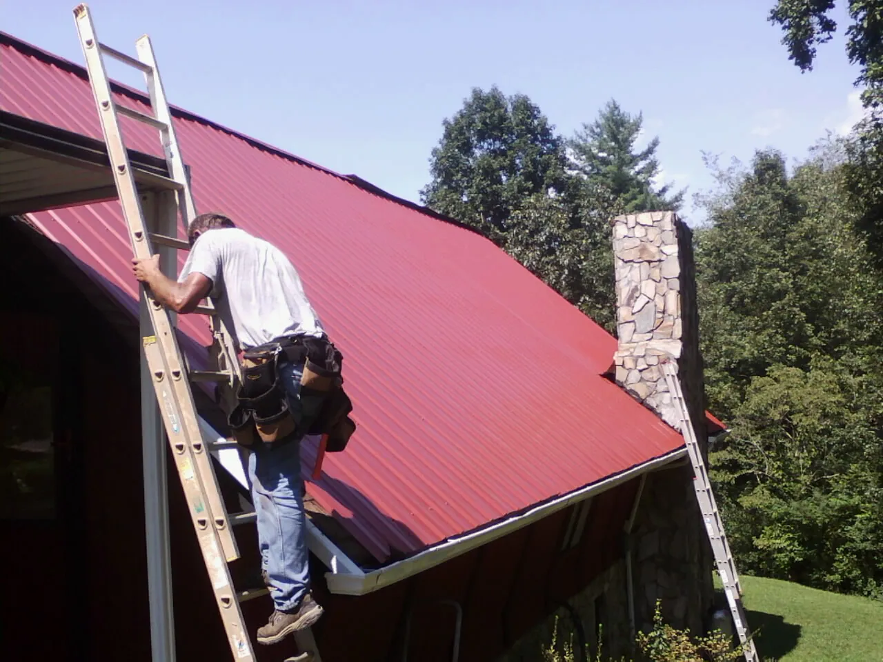 Roofer on a ladder beside a red metal roof and stone chimney — Soffit & Fascia Repair by Roofing Brothers in Hamlet